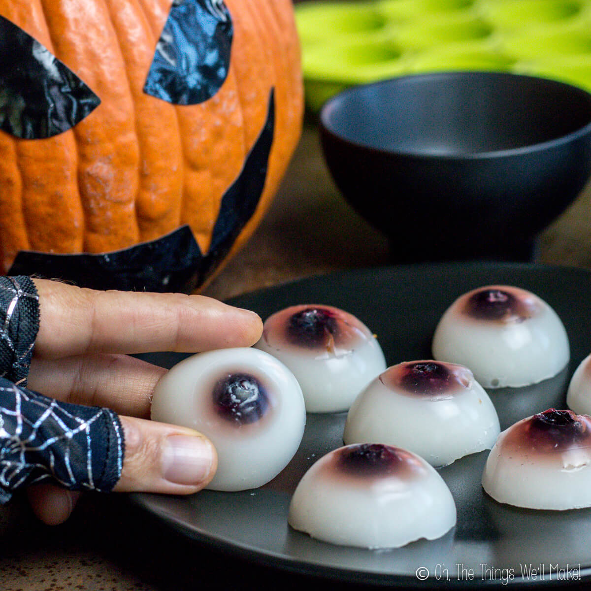 Mano sujetando un ojo de gelatina casero delante de un plato lleno de ojos comestibles para Halloween, con una calabaza decorada al fondo.