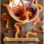 Un cuenco blanco lleno de sanguijuelas de gominola caseras de color naranja y rojo oscuro, rodeadas de más gusanos de gominola esparcidos sobre una superficie oscura.