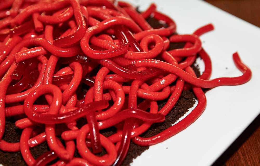 Gusanos de gominola rojos amontonados sobre un plato blanco con una base de migas de chocolate que simulan tierra.