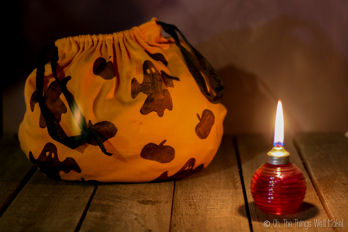 Bolsa de tela naranja para truco o trato, hecha con una camiseta vieja y decorada con fantasmas y calabazas. A su lado hay una pequeña lámpara encendida.