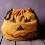 Bolsa naranja con cordón para «truco o trato», decorada con una cara de calabaza de Halloween negra.