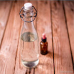 Una botella de cristal transparente llena de vinagre blanco al lado de una botella de aceite esencial sobre una superficie de madera.