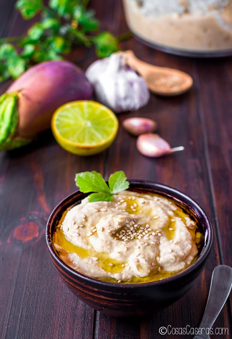  Un cuenco de baba ganoush delante de los ingredientes (una berenjena, medio limón, ajo, y semillas de sesamo)