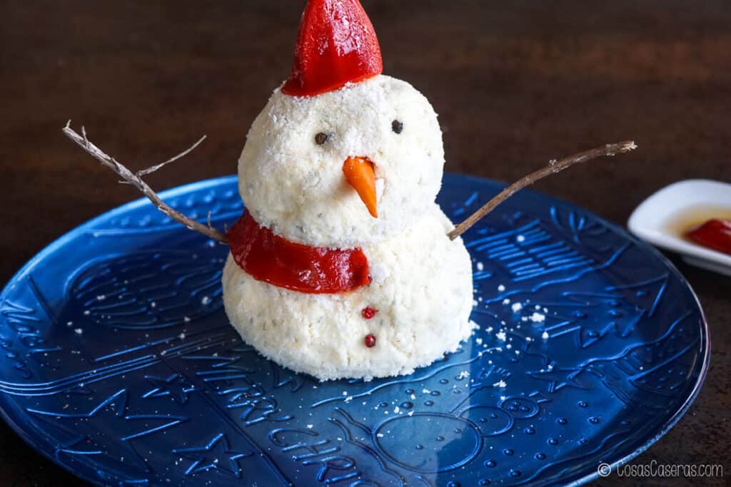 Bola de queso de muñeco de nieve con un sombrero de pimiento rojo asado y una tira de pimiento rojo asado formando la mitad de una bufanda.