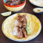 Una tortilla rellena de sabrosas carnitas, yogur griego y guacamole se muestra en un plato. Hay medio aguacate, una rodaja de lima y otros alimentos en el fondo.