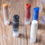 Cinco pequeños envases de maquillaje casero, cada uno de un color diferente, están sobre una superficie de madera. Los colores incluyen negro, blanco, rojo, azul y dorado.