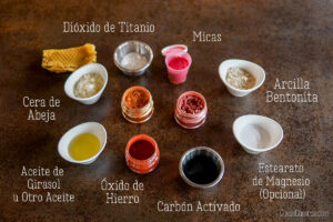 Ingredientes para maquillaje casero: dióxido de titanio, micas, arcilla de bentonita, cera de abejas, aceite de girasol, óxidos de hierro, carbón activado y estearato de magnesio, sobre una superficie marrón oscura.