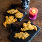 Nuggets de pollo temáticos de Halloween en forma de fantasma y murciélago sobre platos de pizarra, con arañas decorativas, una vela rosa encendida y una calavera plateada en el fondo