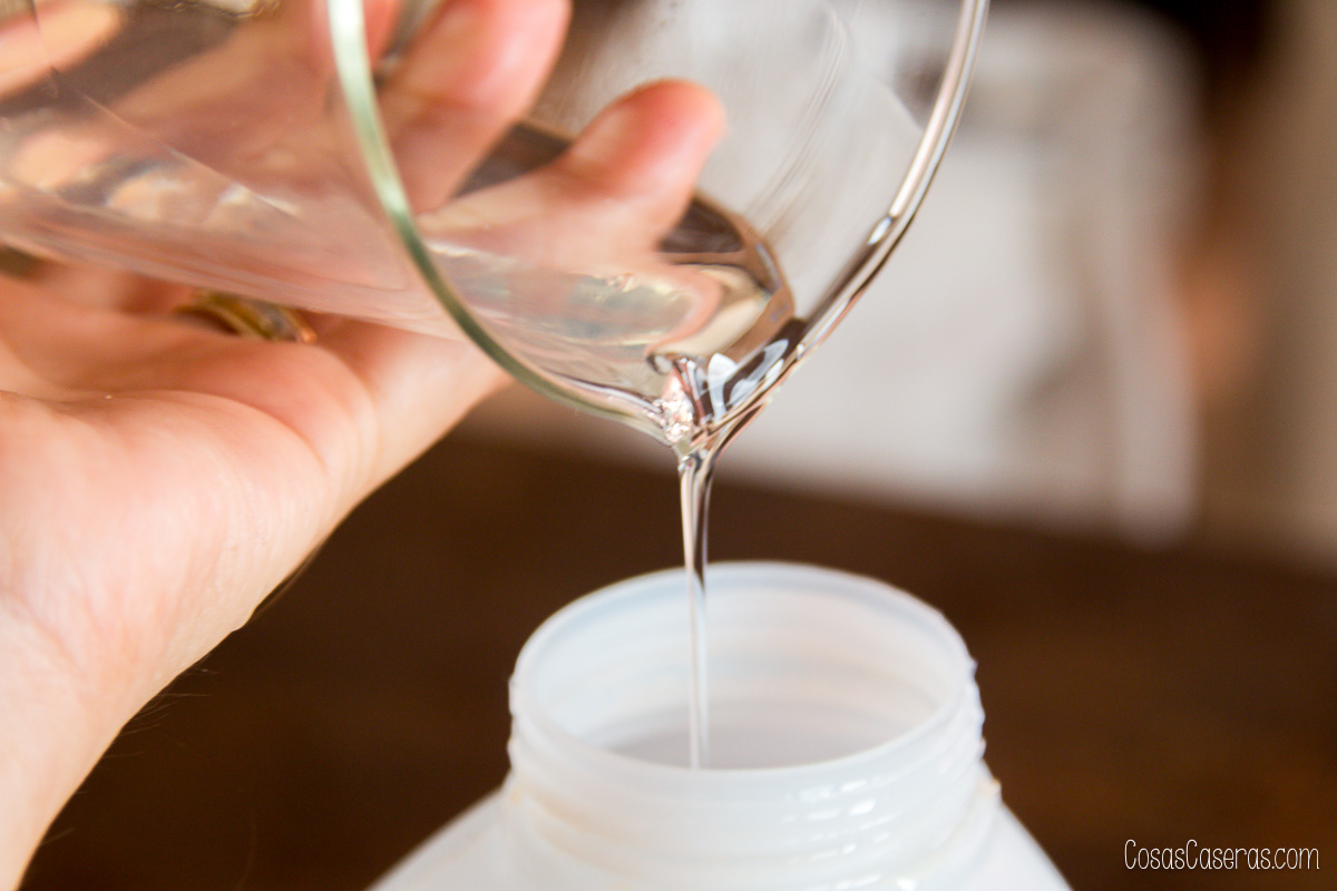 Una mano vierte un líquido claro, probablemente detergente líquido casero, de un recipiente de cristal a una botella de plástico blanca.