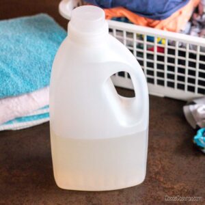 Una botella de detergente de plástico transparente parcialmente llena se encuentra en un mostrador frente a ropa doblada y un cesto de la ropa sucia.