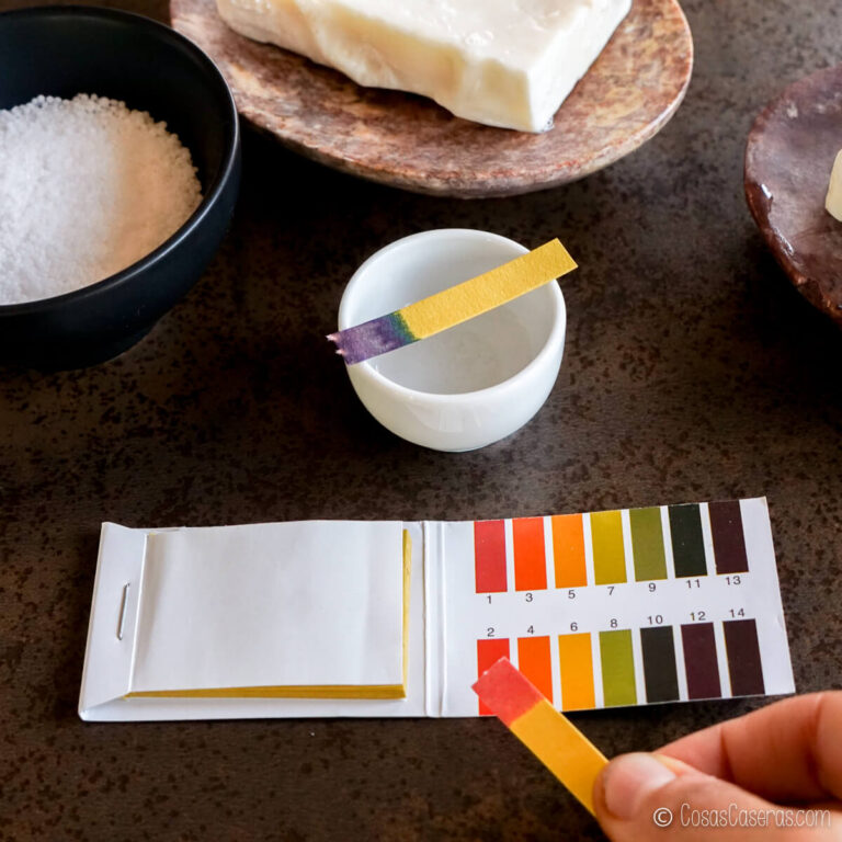 Comparación de una tira reactiva de pH con una tabla de colores, una tira sumergida en un vaso blanco, jabón y sal en cuencos sobre una mesa.