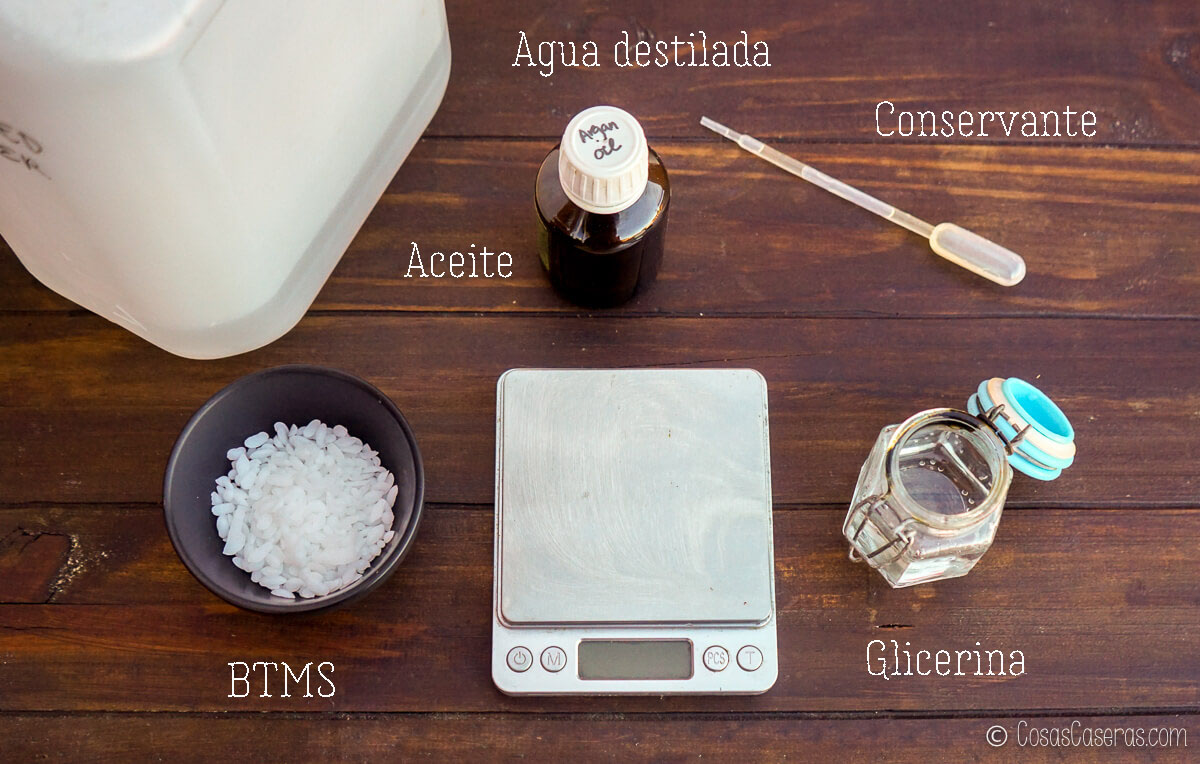 Varios ingredientes etiquetados para una receta cosmética de bricolaje -BTMS, aceite, agua destilada, conservante y glicerina- están dispuestos sobre una superficie de madera con una balanza digital.