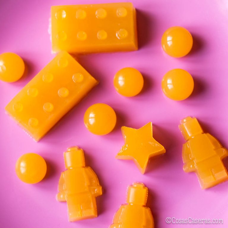Gominolas caseras de color naranja con formas de piezas y figuras LEGO, estrellas y círculos, sobre un fondo rosa.