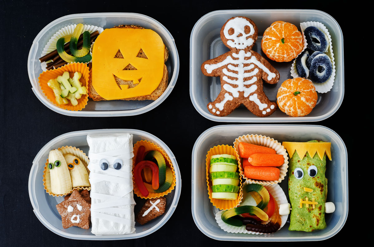 Cuatro tuppers rectangulares con almuerzos inspirados en Halloween, entre los que se incluyen un bocadillo con forma de calabaza de Halloween, una galleta con forma de esqueleto, un tentempié con forma de momia y un wrap de Frankenstein.