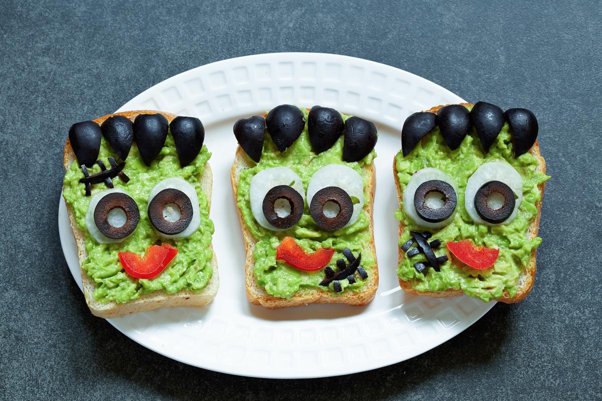 Tres tostadas de aguacate decoradas como el monstruo de Frankenstein, con ojos, pelo y bocas hechos con aceitunas negras y pimiento rojo.