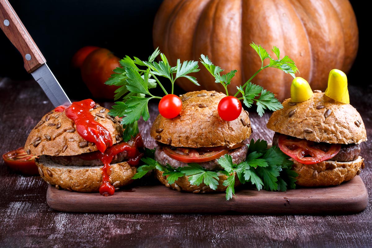 Tres hamburguesas decoradas para Halloween: dos con caras de monstruos y otra con un cuchillo de plástico y kétchup rojo que simula sangre.
