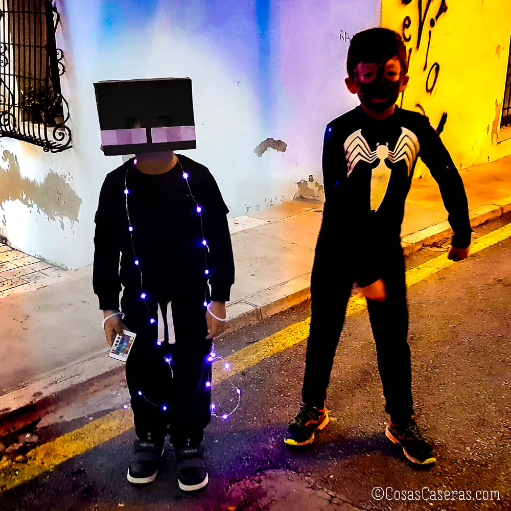 Dos niños con disfraces de Halloween: uno lleva una máscara de caja inspirada en Minecraft con guirnaldas de luces, y el otro lleva un traje de araña al estilo Venom.