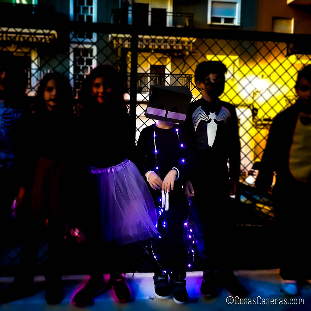 Cuatro niños con disfraces de Halloween de pie delante de una valla metálica, uno en disfraz de enderman con luces moradas.