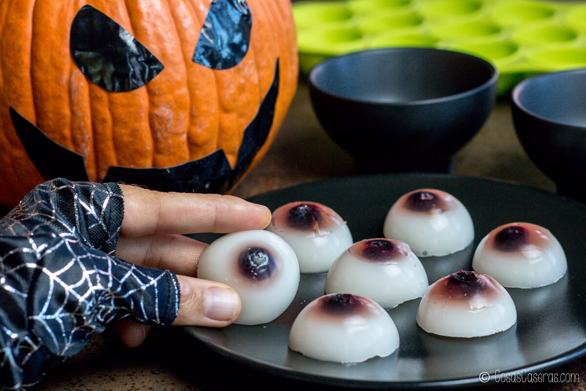 Ojos de gelatina caseros para Halloween, con pupilas de arándanos, sobre un plato junto a una calabaza decorativa.