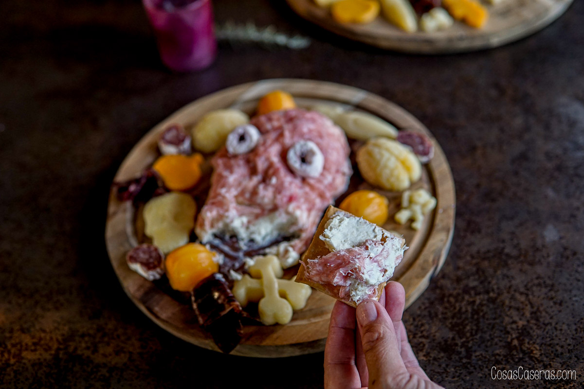 Una mano sostiene una galleta salada con queso de una bola de queso con forma de cara terrorífica, cubierta de embutidos y servida como aperitivo de Halloween.