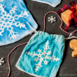 Dos bolsitas de cordón, decoradas con motivos de copos de nieve blancos, acompañadas de pequeños adornos en forma de copo de nieve y una pastilla de jabón envuelta para regalo.