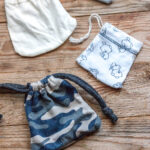 Tres bolsitas de tela con cordón sobre una superficie de madera: una blanca lisa, otra con un estampado de osos y otra con un estampado de camuflaje azul.